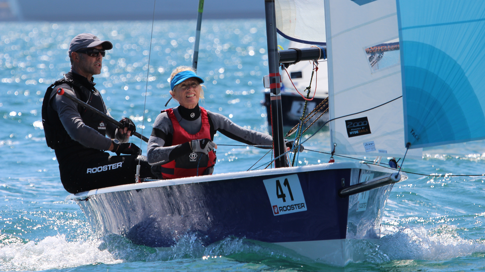 Two adults sailing a Laser 2000 dinghy with blue spinnaker sail at Mylor Sailing School near Falmouth, Cornwall