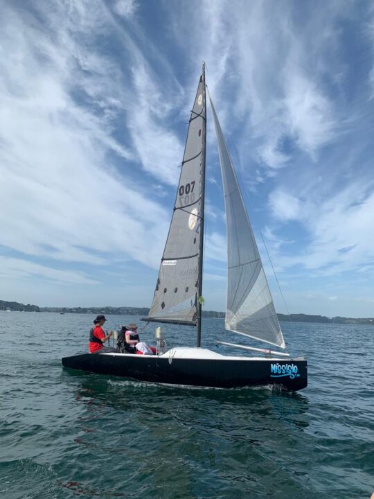 Adapted keelboat sailing with a support seat, a female disabled person being sailed by a female instructor on a sunny day in Mylor Cornwall