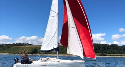 small white sailing yacht with a red downwind spinnaker sail, sailing with 3 people out of Mylor Sailing School Cornwall