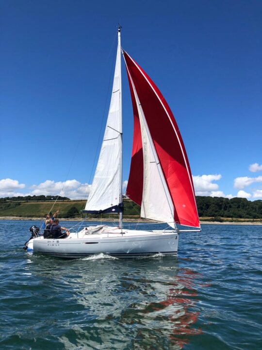 small white sailing yacht with a red downwind spinnaker sail, sailing with 3 people out of Mylor Sailing School Cornwall