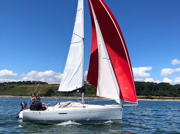 small white sailing yacht with a red downwind spinnaker sail, sailing with 3 people out of Mylor Sailing School Cornwall