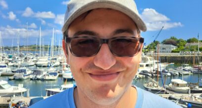 Cam-Underwood-assistant-instructor-young-man-wearing-a-blue-t-shirt-and-cap-smiling-with-a-background-of-boats-on-a-sunny-day-at-Mylor-Sailing-School-Falmouth-Cornwall