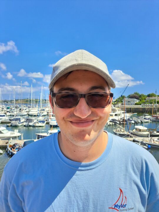 Cam-Underwood-assistant-instructor-young-man-wearing-a-blue-t-shirt-and-cap-smiling-with-a-background-of-boats-on-a-sunny-day-at-Mylor-Sailing-School-Falmouth-Cornwall